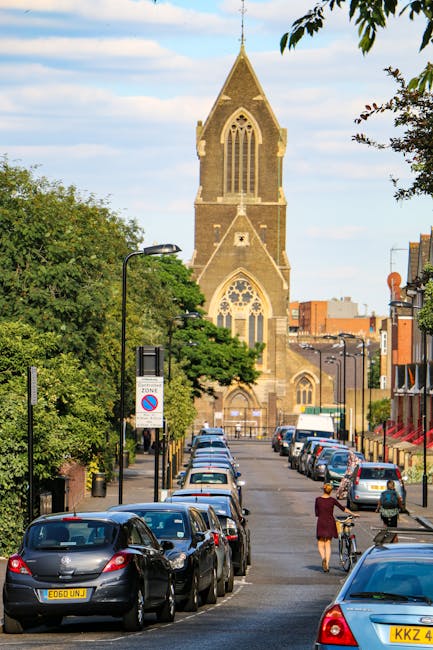 A street scene in Forest Gate lined with parked cars on both sides, including various hatchbacks and sedans, with a woman walking her bicycle and another pedestrian crossing the road. In the background, there is a large historic church with a tall, pointed spire, arched windows, and stone detailing, partially obscured by green trees and foliage. The street is illuminated by black street lamps, and there are several signage posts, including a no parking zone sign, along the pavement. The weather appears clear with a blue sky and some scattered clouds. This scene relates to home relocation and moving logistics, illustrating an area where moving services such as those offered by Man With a Van Forest Gate might operate, especially considering the need to avoid delays in peak times for house removals and furniture transport in E7.