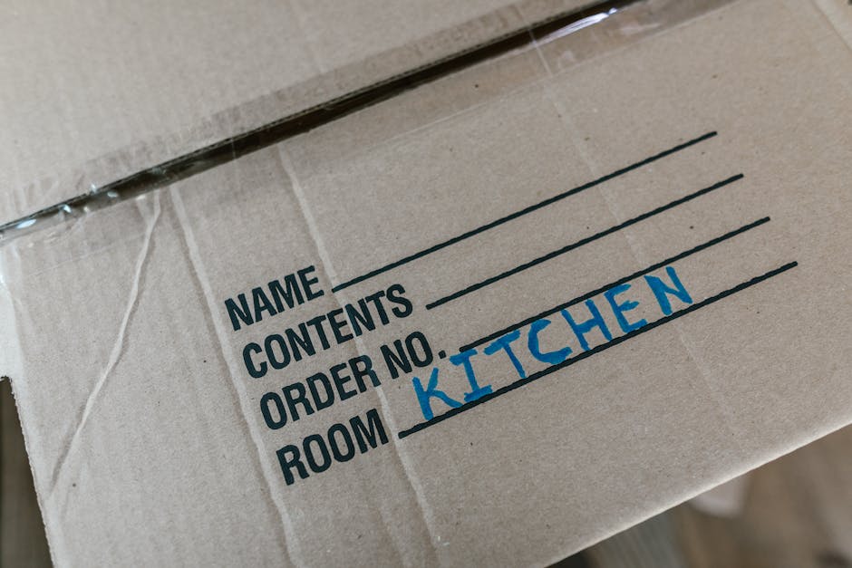 Close-up of a cardboard moving box with printed label sections for 'Name', 'Contents', 'Order No.', and 'Room', with 'KITCHEN' handwritten in blue marker in the 'Room' section. The box appears to be part of a home relocation process, likely being prepared for moving or transport. The background shows additional boxes and packaging materials, indicating an interior setting, possibly a hallway or unpacked room. The scene emphasizes packing activities associated with furniture transport and professional removals, as provided by Man With a Van Forest Gate, highlighting organized packing and careful labeling for efficient home removals or moving services.