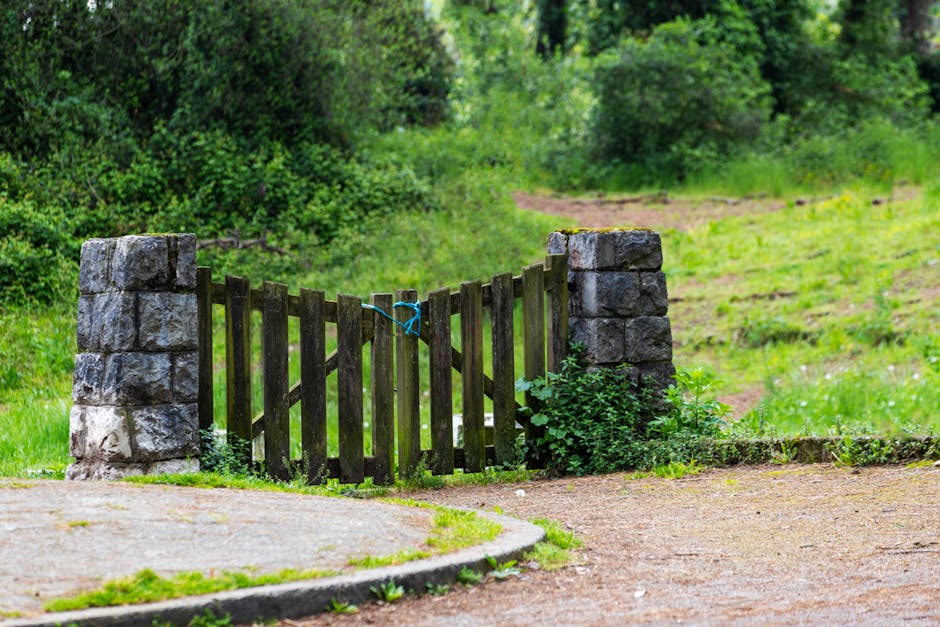 A small wooden gate with vertical slats painted dark brown is positioned between two large stone pillars at the entrance of a garden or pathway. The gate is secured with a blue tie, indicating it may be temporarily closed. Surrounding the gate, there is greenery, including bushes and small plants growing at the base of the stone pillars. In the background, a dirt pathway or track extends through a lush, green landscape with trees and grass, suggesting an area suitable for house removals or home relocation activities. The scene is set outdoors during daylight, with natural light illuminating the foliage and gate, providing a clear view of the entrance typically associated with a rural or semi-rural property. The environment indicates a peaceful, natural setting, and the gate appears to be part of a property boundary, which could be relevant to moving or transport processes managed by companies like Man With a Van Forest Gate.