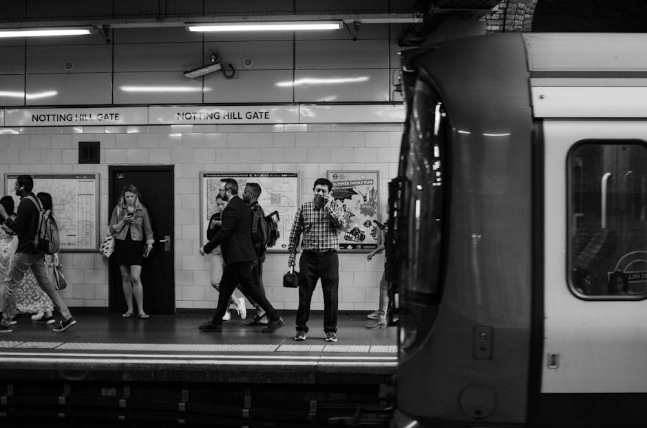 In the foreground of the image, a partially visible train with a sleek, modern exterior is parked at Notting Hill Gate station, with its door open onto the platform. Inside, people are seen waiting and walking along the tiled platform, holding backpacks, shopping bags, and mobile devices. The station’s interior has tiled walls, illuminated by ceiling lights, and features signage indicating the station name. Several passengers, dressed in casual and business attire, are positioned along the platform, some engaged in conversation, others waiting quietly. A man in a checkered shirt with a shoulder bag stands near the train, while others pass by or stand still, showing a typical busy scene of commuters during travel hours. This environment captures the essentials of a London underground station, emphasizing the movement of people during a home relocation or moving process, consistent with services offered by Man With a Van Forest Gate. The lighting and environment reflect the typical underground setting, highlighting the logistical aspects of furniture transport and packing during house removals.