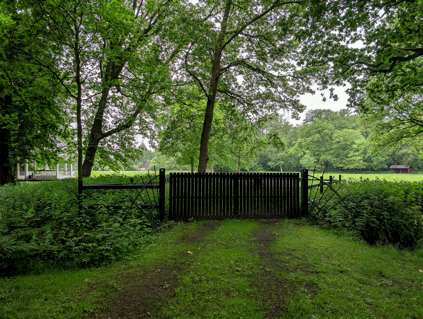 A rural pathway leading through a lush landscape with dense green foliage on both sides, opening up to a bright grassy field beyond a wooden gate. The path is composed of compacted earth and gravel, w