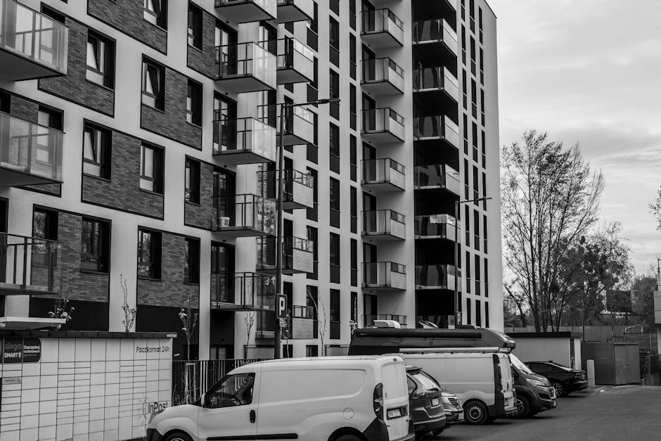 A tall, modern residential building with multiple floors featuring uniform balconies with glass railings, situated in an urban environment. In the foreground, several commercial vehicles, including a