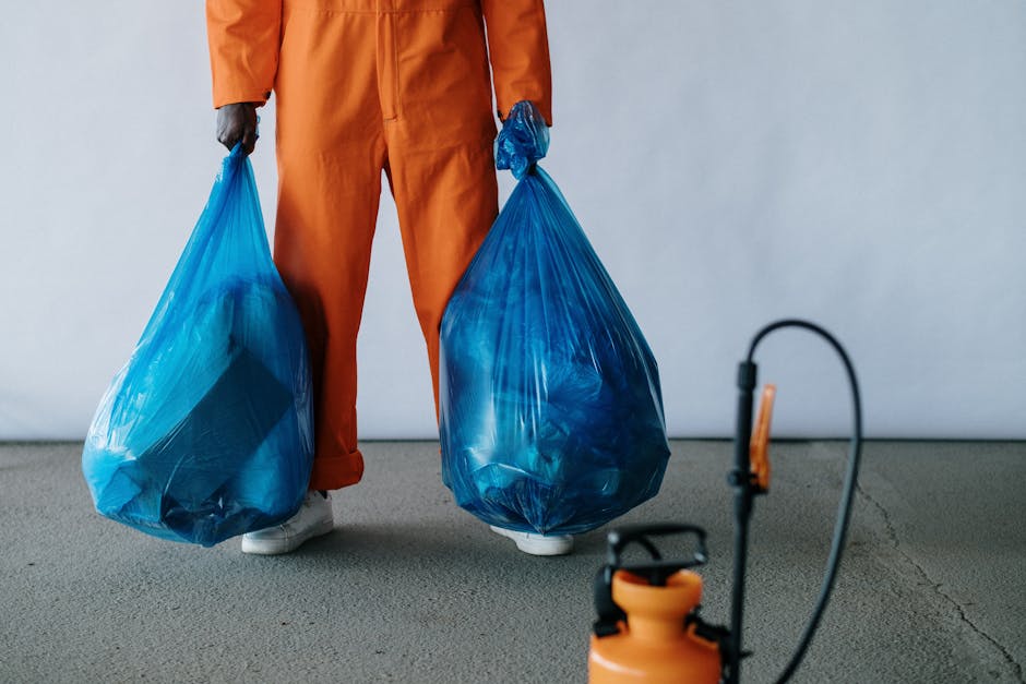 A person dressed in orange overalls is standing inside a property, holding two large blue rubbish bags filled with waste materials, likely for disposal or recycling as part of a home relocation or packing process. The individual is wearing white sneakers and black gloves, with their face not visible. The scene shows the interior space with a plain light-colored wall and a concrete or carpeted floor. In the foreground, a black and orange spray or pest control device is partially visible, suggesting possible cleaning or preparation activities related to a move. The blue bags are filled with miscellaneous waste or packing materials, indicating the clearing out phase typically involved in furniture transport and packing for house removals. This scene illustrates the steps of organizing and waste management during a move, aligning with the services offered by Man With a Van Forest Gate for efficient house removals and right disposal by following local rules, as outlined on the company's page about Forest Gate disposal and recycling rules.