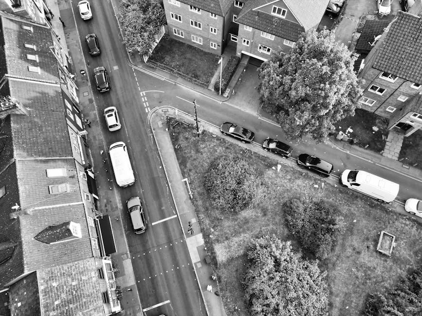 An aerial view of a narrow residential street in Forest Gate, showing several parked cars along the curb and on the adjacent side streets. A long, white van is parked on the right side of the frame, close to a small green space with a few trees and bushes. The van is positioned near the entrance of a property, likely involved in a home relocation or furniture transport process. Surrounding buildings are three to four stories high, with multiple windows and pitched roofs, and a pavement runs along the front of the houses. The scene depicts a typical urban environment suitable for house removals, with the van parked for loading or unloading furniture, boxes, or other household items, and the street is illuminated by daylight, indicating a clear day. This setup is typical for the logistical process of packing, moving, and property clearance, as managed by companies like Man With a Van Forest Gate.
