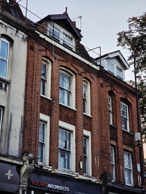 A three-storey brick-built building on Romford Road in Forest Gate, featuring large sash windows on each floor, with two additional floors or roof space visible at the top, including a smaller structure with windows and a pitched roof. The ground floor houses Bolan's Architects, as indicated by a sign, while the upper floors are residential or commercial. The building's facade displays decorative brickwork accents, and part of the roof has metal railings and a small balcony area. Trees are visible to the right side, with foliage partially framing the scene. This image relates to house removals or furniture transport in Forest Gate, with an emphasis on historic building facades typical of urban property relocation projects, often involving careful packing and moving processes by services such as Man With a Van Forest Gate.