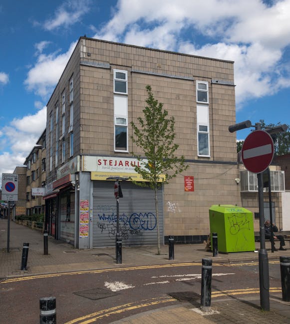 Image of a three-story commercial building on Romford Road in Forest Gate, with a beige brick facade and several white-framed windows. The ground floor features a shuttered storefront with graffiti, and a nearby sign reading 'STEKJARU.' A small tree grows at the corner of the building, and a bright green recycling bin is positioned on the sidewalk. In the foreground, there are multiple black bollards and a pedestrian crossing with double yellow lines on the road. A 'no entry' traffic sign is visible on the right side, and a person is sitting on a bench further along the pavement. The scene is set during daylight with partly cloudy skies, and the overall environment suggests an urban area typical for house removals and furniture transport services, with no visible moving activity in progress.