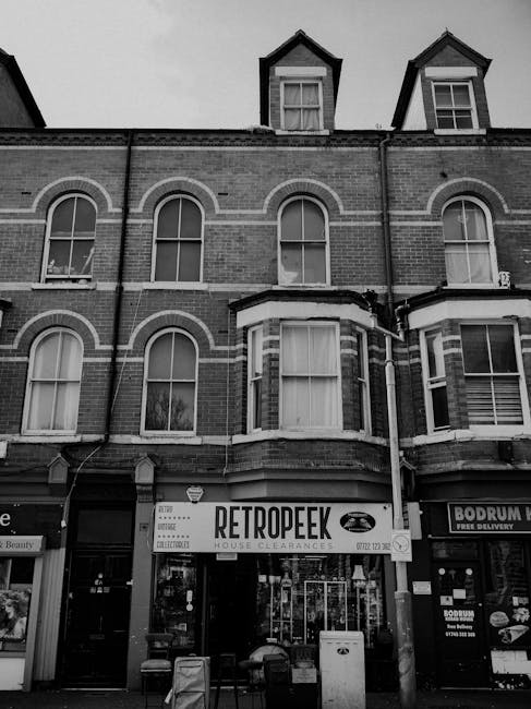 A three-storey brick-built building on Romford Road in Forest Gate, featuring large sash windows on each floor, with two additional floors or roof space visible at the top, including a smaller structure with windows and a pitched roof. The ground floor houses Bolan's Architects, as indicated by a sign, while the upper floors are residential or commercial. The building's facade displays decorative brickwork accents, and part of the roof has metal railings and a small balcony area. Trees are visible to the right side, with foliage partially framing the scene. This image relates to house removals or furniture transport in Forest Gate, with an emphasis on historic building facades typical of urban property relocation projects, often involving careful packing and moving processes by services such as Man With a Van Forest Gate.